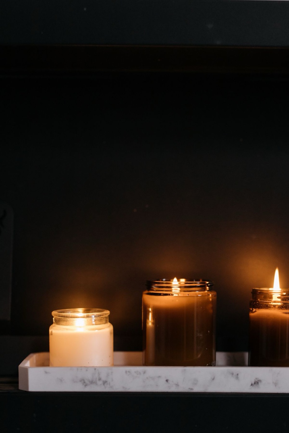 Lit candles in glass jars on a marble tray against a dark wall.