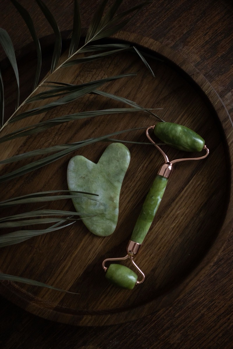A jade gua sha stone and facial roller on a dark wooden tray with a palm leaf.