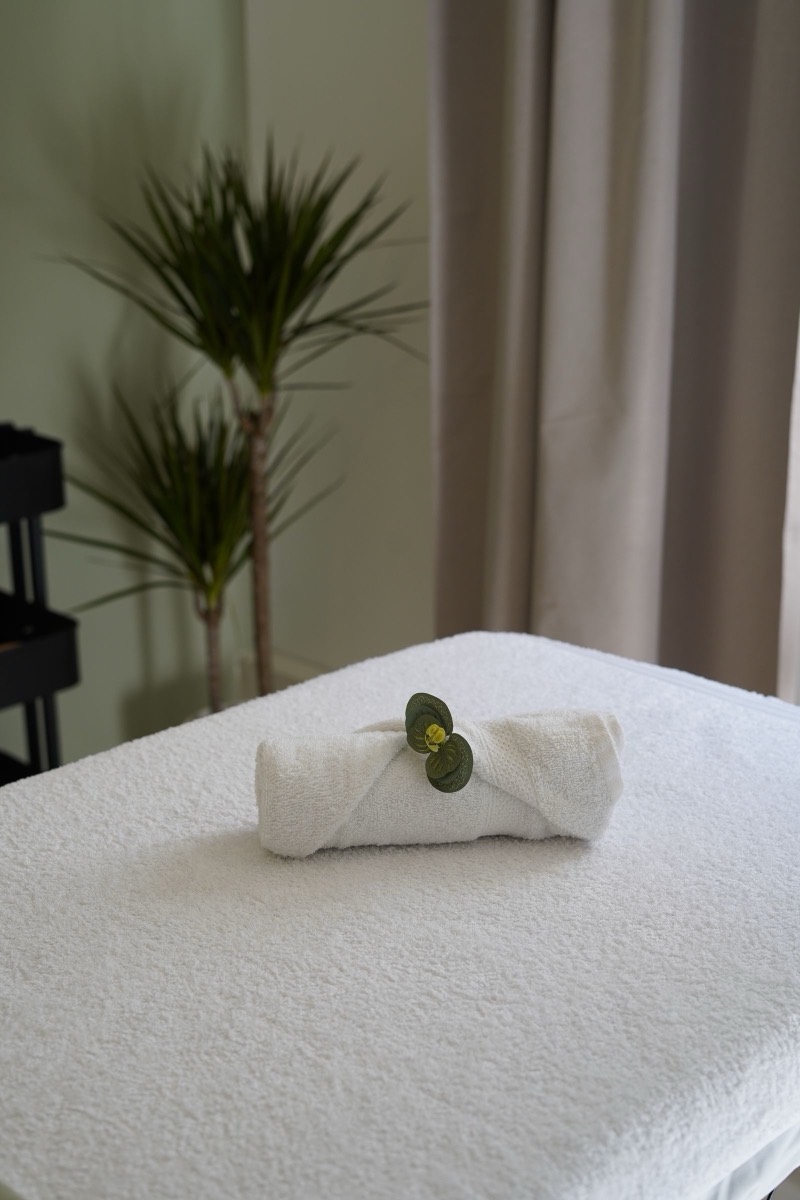 A rolled white towel and smooth stones on a treatment table, a plant behind.