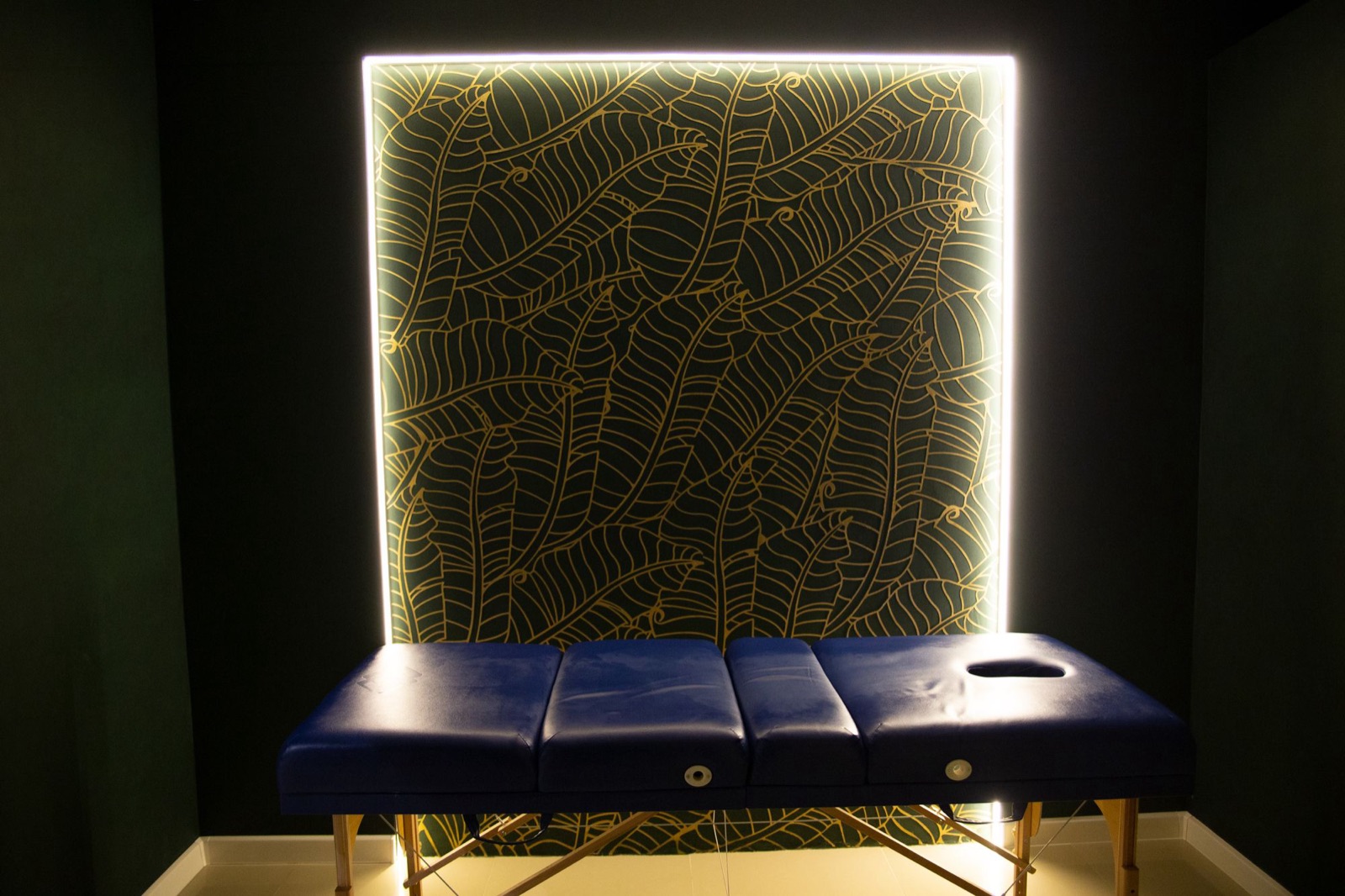A calm treatment space with a backlit gold botanical wall panel and a massage table.